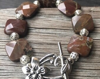 Puede incluir: Un brazalete de piedras preciosas marrón y blanco con un cierre floral plateado. El brazalete está hecho de cuentas cuadradas con acabado facetado.