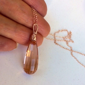 May include: A rose gold necklace with a teardrop-shaped, faceted, light brown gemstone pendant. The pendant is wrapped in rose gold wire.