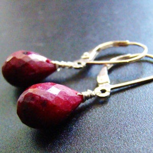 May include: A pair of ruby red teardrop earrings with gold-colored leverback closures. The faceted gemstones are a deep, rich red color. The earrings are displayed on a dark gray surface.