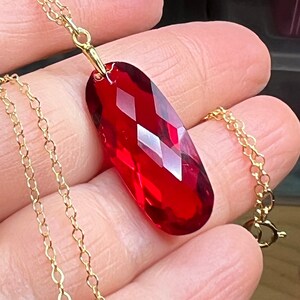 May include: A gold-colored chain necklace with a large, faceted, red gemstone pendant. The pendant is an elongated oval shape with multiple facets that catch the light. The chain is delicate and has a simple clasp.