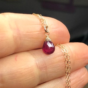 May include: A delicate gold chain necklace with a small, faceted ruby pendant. The ruby is a deep red color and is set in a simple wire wrap.