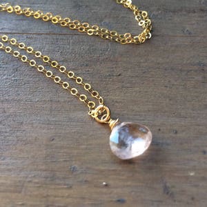 May include: A delicate gold chain necklace with a light pink gemstone pendant. The pendant is a teardrop shape and has a subtle sparkle.