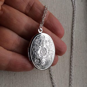 Pode incluir: Um colar de prata com um medalhão oval com um design floral. O medalhão tem uma abertura com dobradiça e uma corrente delicada.