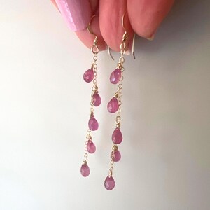 May include: A pair of long, delicate gold earrings with pink gemstone drops. The earrings feature multiple small pink gemstone drops hanging from a thin gold chain.