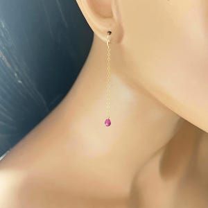 May include: A pair of gold earrings with a small pink gemstone dangling from a delicate chain.
