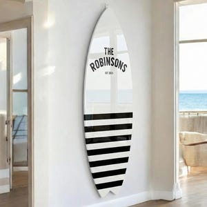 May include: A white surfboard-shaped wall decor with black horizontal stripes on the bottom half. The top half features the text "THE ROBINSONS EST 2023" in black. The decor is mounted on a white wall.