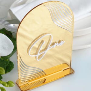 Può includere: Porta numero da tavolo in acrilico dorato con un design a onde e la parola "Uno" in lettere bianche.