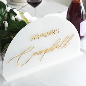 May include: White acrylic half-circle table sign with gold lettering. The sign reads "MR & MRS Campbell".