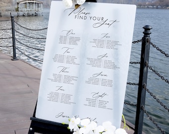 May include: A white acrylic seating chart with a blue watercolor background. The chart has the words "Please Find Your Seat" written in black cursive. The chart is divided into eight sections, each with a number and a list of names. The chart is standing on a black easel.