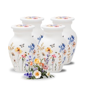 May include: Four white ceramic vases with floral designs. Each vase features a lid with a floral pattern and is adorned with colorful flowers, butterflies, and bees. The vases are arranged together, with a small bouquet of flowers in front.