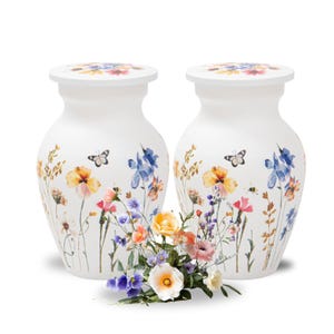 May include: Two white ceramic vases with floral and butterfly designs. The vases have a wide base and a rounded top. The floral design includes yellow, blue, and pink flowers. A small bouquet of flowers sits in front of the vases.