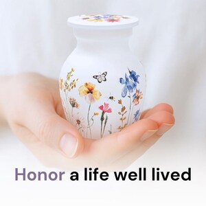 May include: White ceramic urn with a floral design featuring butterflies, bees, and wildflowers in shades of yellow, blue, pink, and green. The urn has a lid and is being held in a hand.
