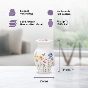 May include: White ceramic urn with a floral design featuring butterflies and wildflowers. The urn is 3 inches tall and 2 inches wide. It comes with a velvet bag and a felt bottom to prevent scratching.