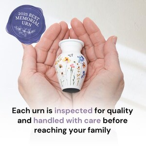 May include: White ceramic urn with a floral design. The urn is being held in two hands. The text "2025 Best Memorial Urn" is on a blue seal.