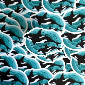 Könnte beinhalten: Nahaufnahme von mehreren gestickten Aufnähern mit einer Mutter und einem Baby-Orca, die in türkisfarbenen Wellen schwimmen. Die Aufnäher sind rund und haben einen weißen Umriss.