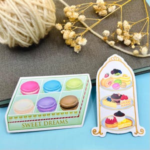 May include: Two stickers: one of a box of colourful macarons and the other of a tiered dessert stand. The macaron box is green with the text "SWEET DREAMS". The dessert stand features various cakes and pastries.