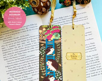 May include: Two bookmarks featuring a hedgehog family theme. The first bookmark shows a ladder with four hedgehogs climbing up. The ladder is surrounded by autumn leaves, berries, and pumpkins. The second bookmark has a beige background with the text "The Hedgehog Family" in a brown circle. Both bookmarks have a gold tassel and bead.