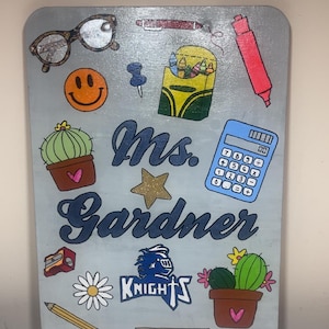 Könnte beinhalten: Rechteckiges, hellgraues Holzschild mit verschiedenen Illustrationen zum Thema Schule. Das Schild zeigt den Namen "Ms. Gardner" in blauer Schrift sowie Bilder von Brillen, einem Smiley, einem Kaktus, einem Taschenrechner und einem Bleistift.