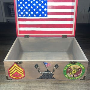 May include: Wooden box with an American flag design on the lid. The box is open, revealing a plain interior. Embellishments include military insignia and a decorative handle. The box is made of wood and has a rustic appearance.
