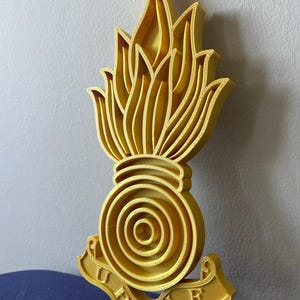 May include: A gold-coloured 3D-printed emblem. It features a flame design above a circular element with concentric rings. A banner at the bottom displays the text "QUE" and "YOU".