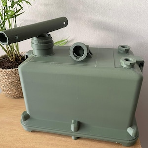May include: A green, rectangular container with a cylindrical top and multiple circular openings. A long, cylindrical piece is attached to the top. The container is sitting on a wooden surface, with a potted plant in the background.