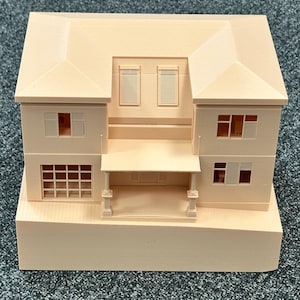Może przedstawiać: Jasny, brzoskwiniowy model miniaturowego domu o dwupiętrowej konstrukcji. Model posiada dwuspadowy dach, liczne okna i ganek wsparty na kolumnach. Dom stoi na prostokątnej podstawie, prezentując detale architektoniczne.