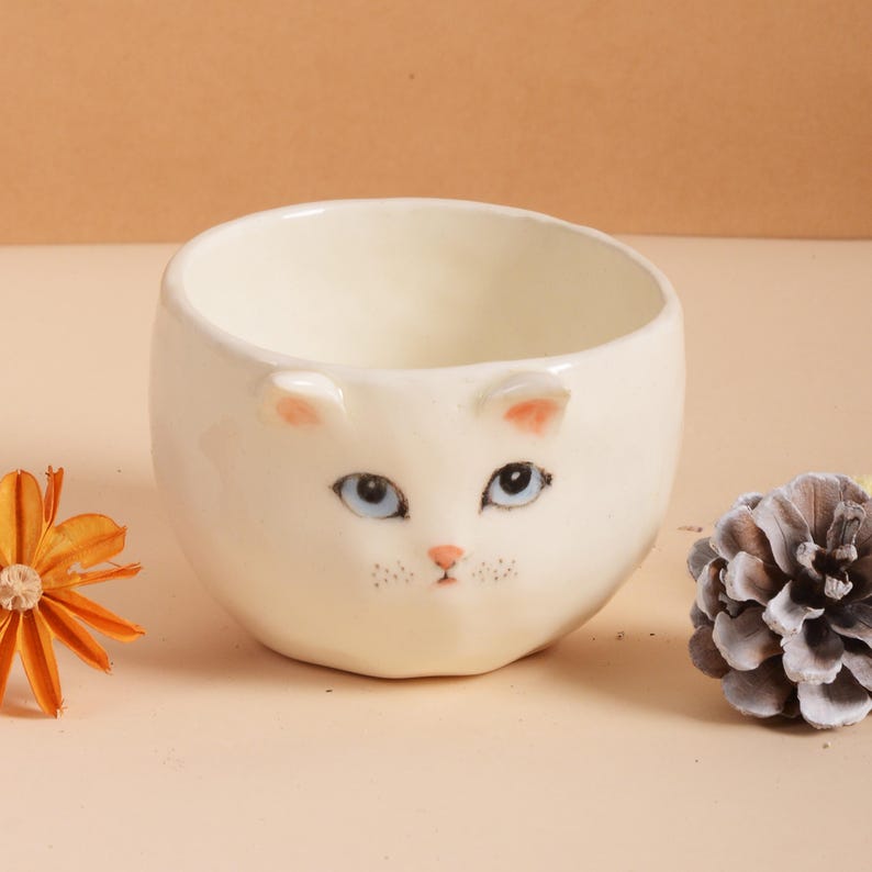 Puede incluir: Un cuenco de cer&aacute;mica blanco con dise&ntilde;o de cara de gato. El cuenco presenta ojos azules, orejas rosas y nariz rosa. El cuenco es redondo y tiene un acabado liso y brillante. Un art&iacute;culo decorativo.