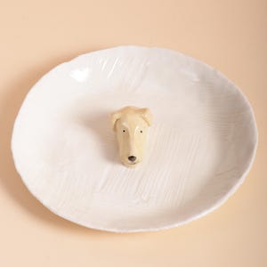 Puede incluir: Un plato de cerámica blanco con una superficie texturizada y una cabeza de perro esculpida en el centro. La cabeza del perro es de color beige claro con detalles negros para los ojos y la nariz. El plato tiene un diseño simple y elegante.