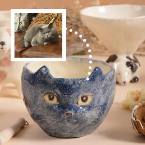 Puede incluir: Un cuenco de cer&aacute;mica azul con un dise&ntilde;o de cara de gato. El cuenco tiene un acabado texturizado y asas con forma de orejas de gato. Los ojos son amarillos y la nariz es marr&oacute;n. Un gato gris est&aacute; en el fondo.