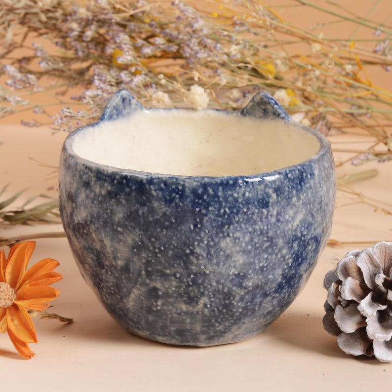 Puede incluir: Un cuenco de cer&aacute;mica azul con un patr&oacute;n moteado y dos peque&ntilde;as protuberancias puntiagudas en forma de oreja. El interior es de color blanco cremoso. El cuenco es probablemente para uso decorativo o para objetos peque&ntilde;os.