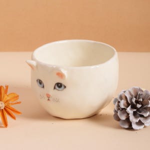Puede incluir: Un cuenco de cer&aacute;mica color crema con forma de gato, con ojos azules, nariz rosa y orejas peque&ntilde;as. El cuenco tiene forma redondeada y es adecuado para plantas peque&ntilde;as o como objeto decorativo. Una pi&ntilde;a y una flor seca est&aacute;n junto al cuenco.