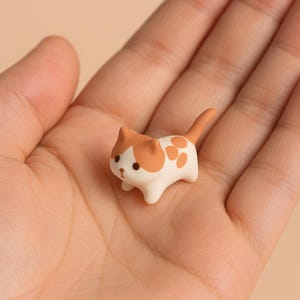 Puede incluir: Pequeña figura de gato de cerámica, con base blanca y manchas naranjas. El gato tiene un cuerpo redondeado, orejas pequeñas y cola. La figura se sostiene en una mano sobre un fondo neutro.