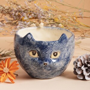 Puede incluir: Un cuenco de cer&aacute;mica azul con un dise&ntilde;o de cara de gato. El cuenco presenta un acabado moteado, con la cara del gato pintada en la parte delantera. Los ojos del gato son amarillos y las orejas puntiagudas. Un art&iacute;culo decorativo &uacute;nico.