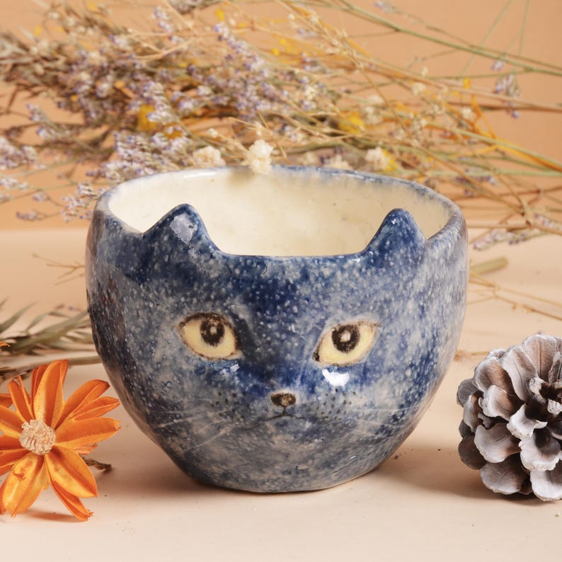 Puede incluir: Un cuenco de cer&aacute;mica azul con un dise&ntilde;o de cara de gato. El cuenco presenta un acabado moteado, con la cara del gato pintada en la parte delantera. Los ojos del gato son amarillos y las orejas puntiagudas. Un art&iacute;culo decorativo &uacute;nico.