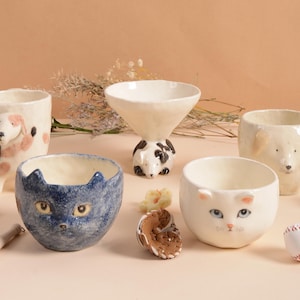 Może przedstawiać: Kolekcja ręcznie robionych ceramicznych misek i kubków, z których każdy ma unikalny wzór twarzy zwierzęcia. Zestaw zawiera elementy z motywem psa, kota i innych zwierząt, prezentujące kapryśną i artystyczną estetykę. Miski są w różnych rozmiarach i kształtach.