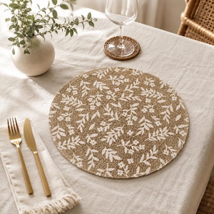 Peut inclure: Un set de table rond tressé en fibres naturelles de couleur beige, orné d'un motif floral et végétal blanc répétitif. Il est posé sur une nappe en lin clair, accompagné d'une fourchette dorée, d'un couteau doré et d'une serviette à franges.