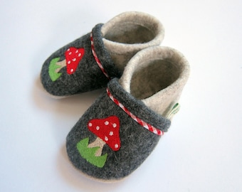 Könnte beinhalten: Ein Paar graue Baby-Filzschuhe mit einem rot-weißen Pilz-Applikation auf der Spitze. Die Schuhe haben eine rot-weiß gestreifte Bordüre um die Öffnung und ein cremefarbenes Innenfutter.