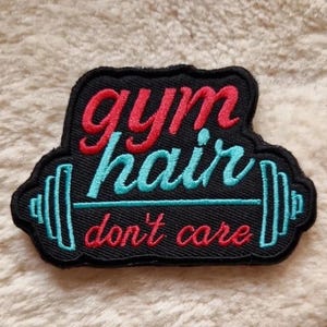 May include: A black embroidered patch with the words "gym hair don't care" in red and teal script. A teal barbell design is below the words. The patch has a black border and is on a beige, textured surface.