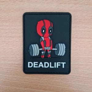 May include: Black patch with a cartoon Deadpool character lifting a barbell. The character is in a red and black suit with a red mask. The word "DEADLIFT" is printed in white below the image.