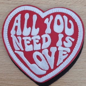 May include: Heart-shaped embroidered patch with the phrase "ALL YOU NEED IS LOVE" in white stitching on a red base. The patch has a black border and is designed for application on clothing or accessories.