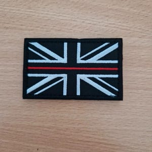 May include: A rectangular black patch displaying a Union Jack design in white and light blue, with a red line across the middle. This patch is likely an emblem or badge, possibly for uniform or apparel.