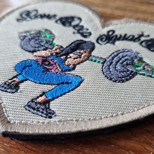 May include: Heart-shaped embroidered patch featuring a weightlifter lifting a barbell. The patch has the text "Love the Squat" in black script. The weightlifter is wearing blue trousers and a pink shirt. The patch has a beige background.