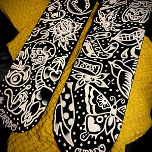 Op de afbeelding: Twee zwarte skateboards met witte tattoo-achtige ontwerpen. Eén heeft een schedel, roos en kers, terwijl de andere een vlinder, hart en het woord "LUCK" heeft. Het woord "CURADO" staat onderaan elk board.