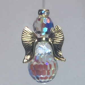May include: A decorative angel ornament with iridescent crystal-like body and head, gold-colored wings, and a small gold-colored halo. The ornament is suspended by a thin, clear string.