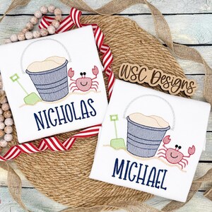 May include: Two white t-shirts with embroidered images of a bucket and shovel, a crab, and the names "Nicholas" and "Michael".