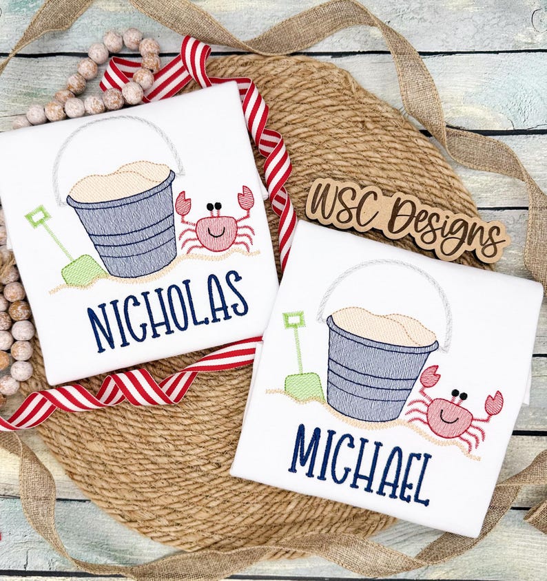 May include: Two white t-shirts with embroidered images of a bucket and shovel, a crab, and the names "Nicholas" and "Michael".