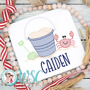 May include: Embroidered design of a blue bucket with sand, a green shovel, and a red crab with a smiling face. The name "CAIDEN" is embroidered below the design.