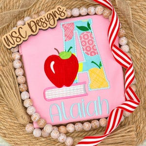 May include: Pink t-shirt with a large embroidered letter "A" decorated with fruit and floral patterns. A red apple and books are embroidered below the letter. The name "Alaiah" is embroidered in light blue.
