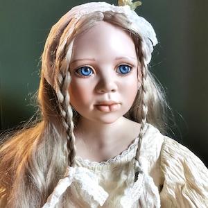 May include: A porcelain doll with long, blonde hair styled in plaits. The doll has bright blue eyes, rosy cheeks, and a delicate expression. She wears a cream-coloured dress and a white lace bonnet with a small floral accent. The doll is a collectible item.
