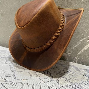 May include: A brown leather cowboy hat with a braided band around the crown. The hat has a wide brim and a slightly curved crown. The leather has a distressed look, giving it a vintage appearance.
